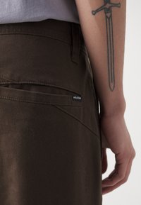 Braune Volcom-Hosen haben eine glatte Textur und eine Gesäßtasche mit einem schwarzen Logo-Tag. Ein tätowierter Arm lehnt gegen den Stoff.