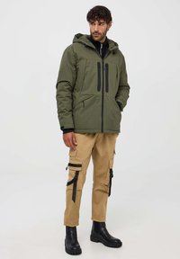 Giacca imbottita verde oliva con cappuccio, chiusura frontale con zip, due tasche sul petto con cerniere, indossata con pantaloni cargo beige e stivaletti neri.