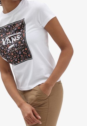 Osoba ubrana w białą koszulkę Vans z kwiatowym logo i beżowe spodnie, stojąca z jedną ręką w kieszeni, a drugą swobodnie opuszczoną wzdłuż ciała.