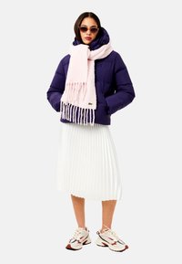 Jachetă pufosă purpurie, eșarfă cu franjuri roz deschis, fustă plisată albă și pantofi sport. Ochelarii de soare completează aspectul modern.