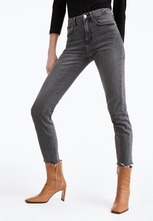 Žena oblečená ve šedých úzkých džínách s vysokým pasem a třepícími se lemy a v hnědých kotníkových botách na podpatku, stojící s rozkročenýma nohama.