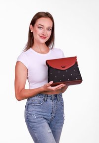 Černá a červená kabelka s hladkým koženým klopou, tělem z látky s puntíky a akcentem loga. Modelka drží kabelku v dobře osvětleném prostředí.