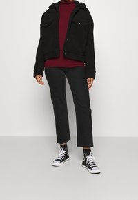 Svart fleecejacka med två bröstfickor, bärs över en vinröd polotröja, i kombination med svarta raka jeans och svarta högskaftade sneakers.