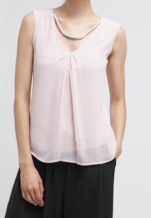 Vrouw draagt een mouwloze lichtroze blouse met plooidetail aan de voorkant en een zilveren gebogen ketting, gecombineerd met een zwarte broek.