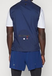 Marineblaues ärmelloses Vest mit glatter Textur, ausgestattet mit einer Rückentasche mit Reißverschluss und rotem Ziehband. Über einem weißen Hemd getragen.