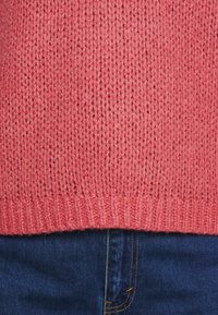 Pletený svetr v korálovém odstínu s volnou vazbou a žebrovaným lemem, doplněný tmavým dením v pase.