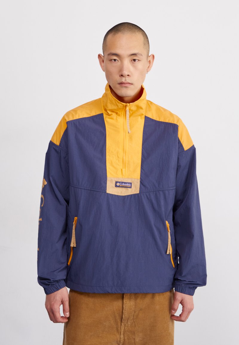 Chaqueta cortavientos en azul marino y amarillo con cremallera de medio largo en la parte delantera, puños elásticos y bolsillos laterales. Presenta un parche de logo en el pecho.