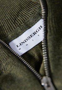 Olivgrön stickad tröja med dragkedja, med en vit etikett inskriven "LINDBERGH WHITE" i svart text. Närbild av den texturerade tyget.