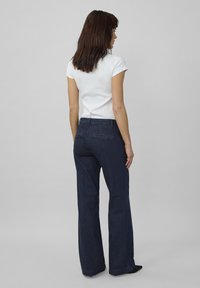 Een vrouw staat met haar rug naar de camera, gekleed in een witte kortekoope top en donkerblauwe flare jeans tegen een grijze achtergrond.