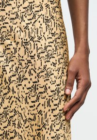 Σκούρο δέρμα χεριού που ακουμπά δίπλα σε μπεζ ύφασμα με ακανόνιστο μαύρο γεωμετρικό σχέδιο, κοντινή λήψη στην περιοχή του χεριού και της μέσης.