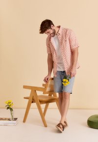 Korte mouwen shirt met rood geometrisch patroon, wit T-shirt, spijkerbroek shorts en bruine sandalen. Houten stoel en gele bloemen in een vaas in de buurt.