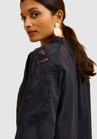 Blusa in stoffa nera con ricamo floreale e inserti trasparenti sulle maniche. Presenta un colletto arricciato e maniche lunghe.