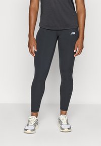 Leggings de sport noirs avec une coupe ajustée, en matériau extensible, présentent un petit logo sur la hanche et sont associés à des baskets grises.