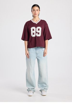 Maroon oversized jersey with white stripes and number 89 on the front. Paired with light blue wide-leg jeans and white sneakers.