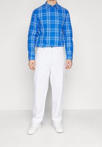 Camicia a quadri blu con linee bianche, maniche lunghe. Abbinata a pantaloni bianchi larghi e sneakers bianche su uno sfondo neutro.