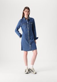 Vestido camiseiro de denim em azul escuro com mangas longas, abertura frontal com botões e bolsos no peito, apresentando rachas laterais e uma barra ligeiramente arredondada.