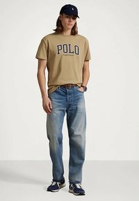 Νεαρός άνδρας που φοράει μπεζ μπλουζάκι Polo Ralph Lauren, μπλε τζιν, ναυτικές αθλητικές παπούτσια και ναυτική καπέλο, στέκεται με το ένα χέρι στην τσέπη.