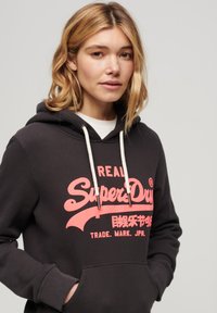 Felpa grigia scura con un grande logo rosa "SuperDry", cordini color crema, tasca a marsupio e polsini a coste. Tessuto in misto cotone.