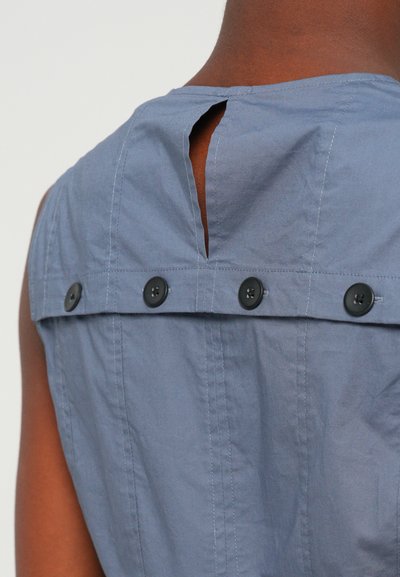 Mėlyna be rankovių palaidinė su skyle nugaroje, penki juodi mygtukai per petnešas ir tekstūruota medvilninė audinys.