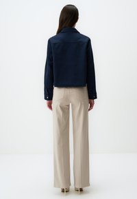Marineblaues Cropped-Jacket mit großem Kragen, kombiniert mit hochtaillierten beige-weiten Hosen. Glatter Stoff mit sichtbaren Nahtdetails an der Rückseite.