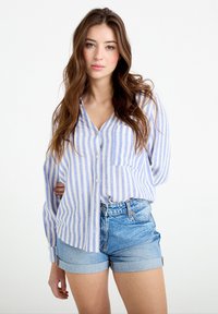 Joven con cabello largo y castaño, vestida con una camisa de botones a rayas azul y blanca y shorts de mezclilla arremangados, de pie frente a un fondo blanco.