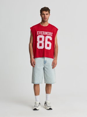 Uomo che indossa una maglia sportiva senza maniche rossa con la scritta "EVERMORE 86", pantaloncini corti di jeans chiari fino al ginocchio, calzini bianchi e sneakers grigie, in piedi di fronte.