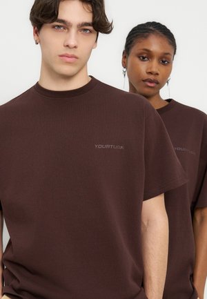 Jeune homme et femme portant des t-shirts brun foncé "YOURTURN", debout côte à côte avec des expressions neutres sur un fond clair uni.