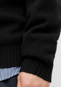 Sort strikket sweater med ribbede manchetter, der fremviser en fin tekstur. Lagd ovenpå en blå og hvid stribet skjorte.