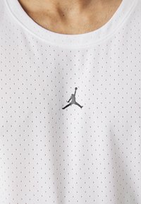 Bílý sportovní tílko s malými perforacemi, na hrudi s černým logem Jumpman. Měkká látka s žebrovaným výstřihem.