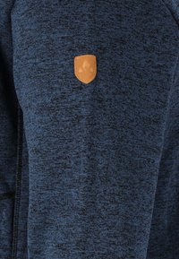 Tummansininen huppari, jossa on teksturoitu kangas, ja vasemmassa yläkulmassa tan-värinen nahkapatch-logo sekä täysipitkä vetoketju.