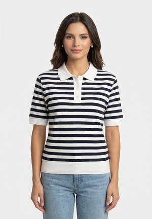 Jeune femme aux cheveux foncés portant des boucles d'oreilles en perles, un polo à manches courtes blanc et rayé bleu marine, et un jean bleu clair, debout, droite.