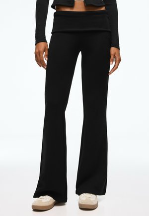 Personne portant un pantalon noir taille haute évasé, des baskets beiges et un haut noir à manches longues montrant le ventre, debout sur fond blanc.