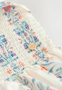 Detailný záber na krémovú látku s modrými a ružovými kvetinovými vzormi, s malým okrúhlym zlatým gombíkom s reliéfnym motívom.