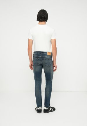 Homme debout, dos tourné, portant un t-shirt blanc, un jean slim bleu foncé délavé et des baskets noires avec des bandes blanches.
