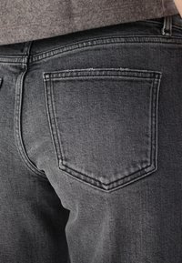 Gros plan d'une personne portant un jean en denim noir délavé montrant la poche arrière et une partie d'un pull gris.