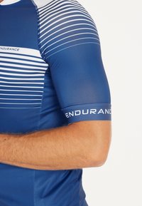 Sininen lyhythihainen urheilupaita valkoisilla vaakasuorilla raidoilla, jossa on "ENDURANCE"-logo hihassa, valmistettu sileästä ja joustavasta materiaalista.