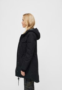 Svart parka-jakke med hette, har frontlommer, glidelås foran og innsnevring i midjen. Struktureret stoff med matt finish.
