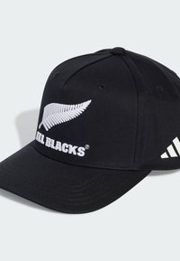Cappello da baseball nero in tessuto, con un logo in ricamo bianco a forma di felce e il testo "ALL BLACKS" sulla parte frontale, con tre strisce bianche sul lato.