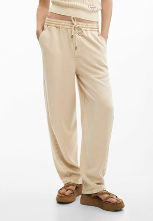 Personne portant un pantalon beige à cordon de serrage et des sandales à plateforme marron avec des brides, debout avec les mains dans les poches sur un fond blanc.