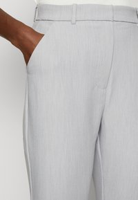 Světle šedé kalhoty vyrobené z jemně texturované tkaniny, s bočními kapsami a přizpůsobeným střihem. Podrobnosti švů jsou viditelné podél okrajů.