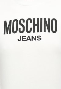 Λευκό ύφασμα με μεγάλα μαύρα γράμματα που γράφουν "MOSCHINO" πάνω από μικρότερα μαύρα γράμματα "JEANS" στο κέντρο της επιφάνειας.