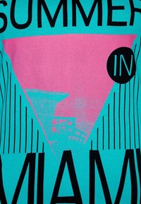 Camiseta de algodón turquesa con un gran gráfico de triángulo rosa y texto en negro "VERANO EN MIAMI" con acentos de rayas verticales en negro.