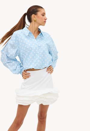 Femme portant une chemise bleue à pois à manches longues et une jupe blanche à volants superposés, debout les mains sur les hanches, cheveux attachés en queue de cheval, de profil.