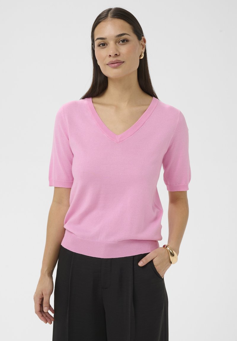 Mujer de cabello largo y oscuro, que lleva un suéter rosa de manga corta y cuello en V, y pantalones negros, complementada con pendientes de aro dorados y una pulsera de oro.