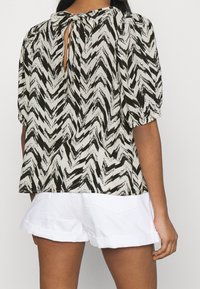 Blusa a zigzag bianca e nera con chiusura a cravatta al collo e maniche a sbuffo, abbinata a pantaloni corti bianchi.