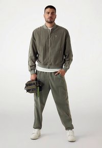 Lichtgewicht olijfgroene bomberjack met ritssluiting, geribbelde manchetten, bijpassende broek en een zwart-groene utilitytas. Witte sneakers.