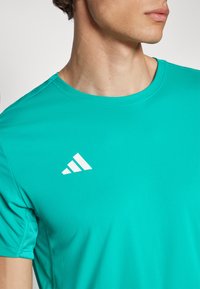 Tirkizna sportska majica s okruglim izrezom, s bijelim logotipom od tri pruge na gornjoj lijevoj strani i mrežastim detaljima na rukavima.
