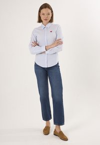 Camicia a righe blu chiaro e bianca con bottoni, caratterizzata da un piccolo logo a forma di cuore rosso sul petto, abbinata a jeans dritti di denim e mocassini marroni.