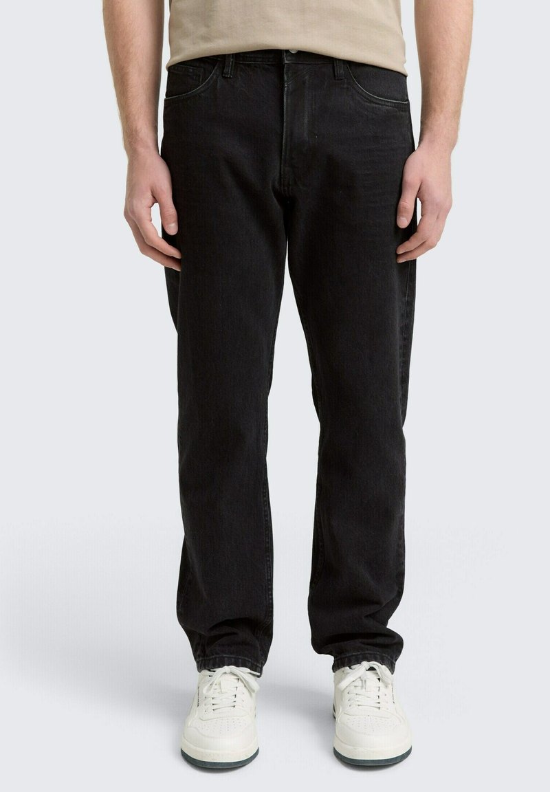 Mann iført rette svarte jeans og hvite sneakers som står mot en lys bakgrunn med hendene avslappet langs siden.