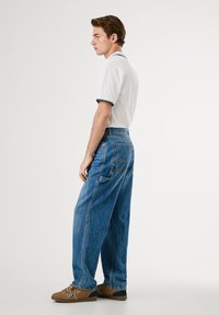 Hvit polo-skjorte med marineblå detaljer, avslappet passform lys blå denimbukser og brune sko med en mørkere aksent. Sidesyn, stående posisjon.
